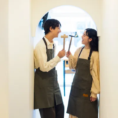 アーチ型の通路でエプロンを着けた男女が向かい合い、木槌を持ちながら笑顔で見つめ合っている。