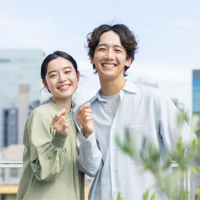 都会の屋上で、男女が指でハートマークを作り、カメラに笑顔を向けている。