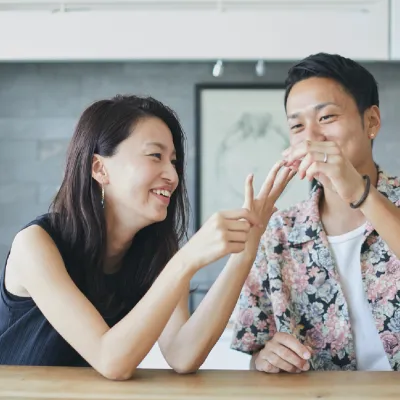 室内でテーブル越しに座る男女が、指でリングをはめる仕草をしながら笑顔を交わしている。