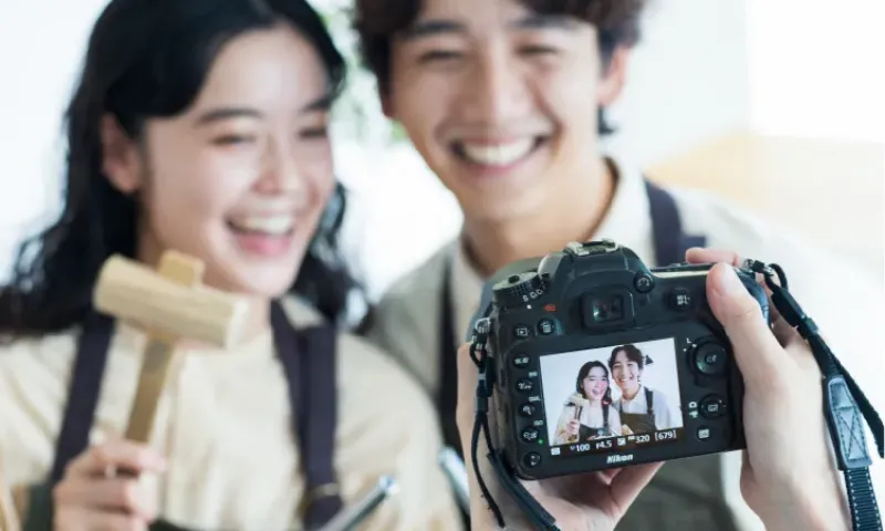 エプロン姿の男女が笑顔で寄り添い、手前のカメラでその様子を撮影している。女性は木槌を持っている。