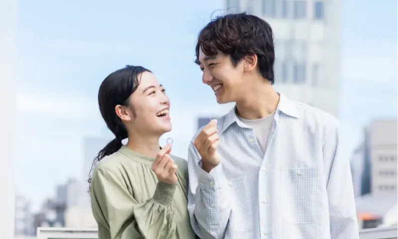 屋外で、男女が小さな輪を手に持ち、見つめ合いながら笑顔を交わしている。背景には高層ビルが見える。